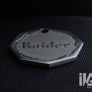 May include: A silver octagonal tag with the word "Raider" engraved on it.