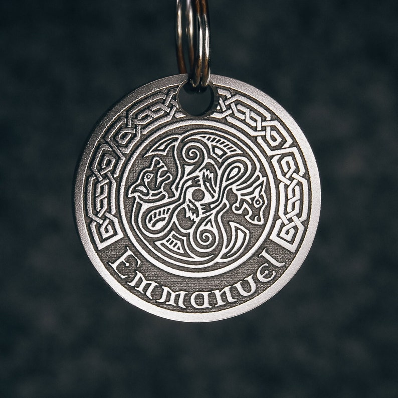 Puede incluir: Un llavero de metal plateado con un dise&ntilde;o de nudo celta y la palabra "Emmanuel" inscrita alrededor del borde. El centro del llavero presenta un dise&ntilde;o detallado de tres criaturas entrelazadas.