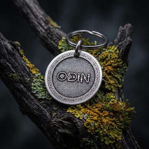 Puede incluir: Una etiqueta circular de metal plateado para llavero con un centro texturizado donde aparece grabada la palabra ODIN en una fuente rúnica estilizada. El borde exterior del disco está decorado con un intrincado patrón de trenzado.