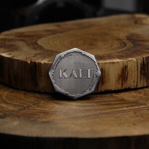 May include: A silver-colored, octagonal-shaped metal token with the word "KALI" engraved on it. The token is sitting on a wooden surface.