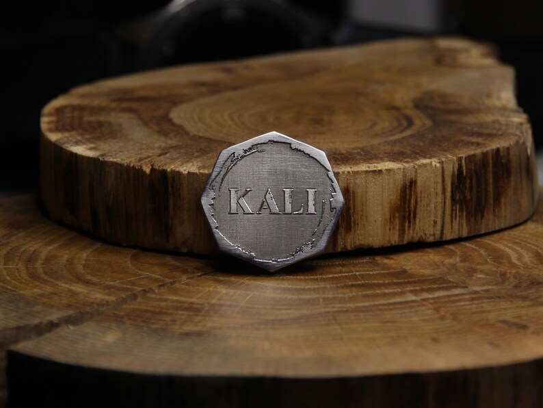 May include: A silver-colored, octagonal-shaped metal token with the word "KALI" engraved on it. The token is sitting on a wooden surface.