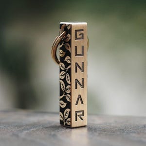 Puede incluir: Un llavero de metal dorado con un diseño negro y dorado. El llavero tiene un patrón de hojas en un lado y el nombre "GUNNAR" grabado en el otro.