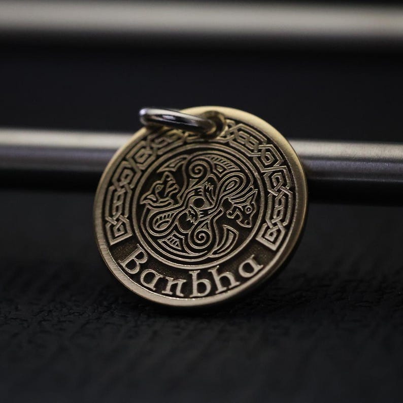 A close-up of a round, golden metal medallion with Celtic-style patterns, a central design of mythical animals, and the word "Banbha" engraved at the bottom. It hangs from a loop on a metal rod against a dark, blurred background.