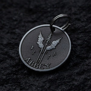 Könnte beinhalten: Ein schwarzer Metall-Hundeanhänger mit einem silbernen Flügeldesign und dem Wort "ABBEY" am Rand.