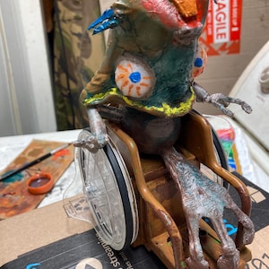 May include: A hand-painted creature figurine with a large, textured head, prominent white eyes with orange irises and blue pupils, and a brown body. The creature is seated in a brown plastic wheelchair with clear wheels and a long, textured tail.