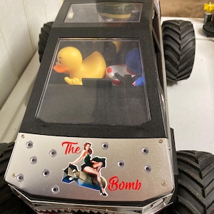 May include: A black monster truck toy with large tires. The front features a silver panel with the text "The Bomb" and a pin-up girl graphic. Inside the truck are a yellow rubber duck and other small toys. A green bomb-shaped object sits on top.