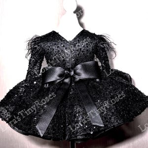 Könnte beinhalten: Schwarzes Paillettenkleid mit langen Ärmeln und einer großen Satinschleife in der Taille. Das Kleid hat Federn an den Schultern und einen V-Ausschnitt. Der Text "Lora's Tiny Roses" ist sichtbar.