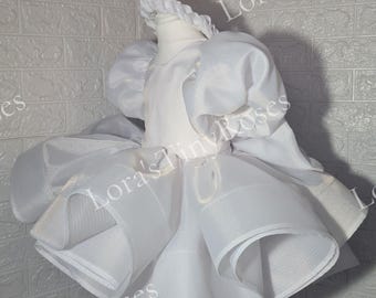 May include: A white dress with a layered skirt and puffy sleeves, displayed on a mannequin. The dress has a matching headband. The fabric appears to be a lightweight, possibly organza or similar, creating a delicate, airy look.