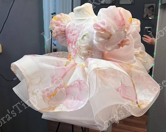 Op de afbeelding: Een witte tule jurk met roze bloemenprint en pofmouwen. De jurk staat op een witte standaard.