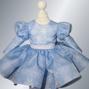 May include: A light blue dress with a snowflake pattern, featuring long sleeves and a full skirt. The dress has a white waistband and is displayed on a mannequin. The dress is designed for a child.