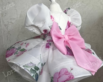 Op de afbeelding: Een witte jurk met een bloemenpatroon van roze rozen en groene bladeren, ontworpen met volumineuze pofmouwen en een grote, opvallende roze strik in het midden. Het kledingstuk wordt getoond op een paspop en heeft een wijde rok.