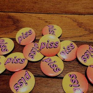 May include: A collection of 11 round yellow and orange buttons with the word "piss" written in pink letters on each button.