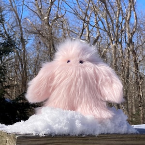May include: A fluffy, light pink creature with small, purple eyes. The plush toy has a shaggy appearance and is sitting on a bed of snow. The background features bare trees and a clear blue sky, suggesting a winter scene.