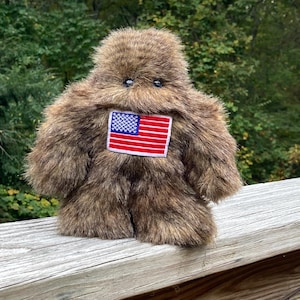 May include: A brown plush toy in the shape of a Bigfoot-like creature. It has small black eyes and a patch of the American flag on its chest. The toy is made of a shaggy, fur-like material.