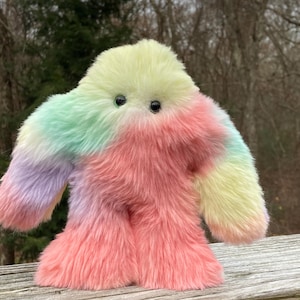 May include: A plush, colorful creature with a soft, furry texture. The toy features a pastel color palette of yellow, pink, green, and purple. It has two small black eyes and a rounded body with arms and legs.