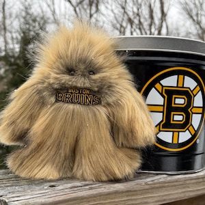 Peut inclure: Une créature en peluche, marron clair et touffue, avec le texte "BOSTON BRUINS" sur la poitrine, à côté d'une boîte en métal noire et jaune avec le logo des Bruins. La créature a une texture poilue. La boîte affiche le logo des Bruins.