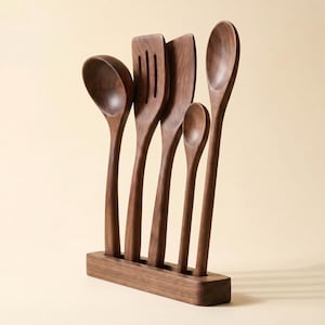 May include: A set of five wooden kitchen utensils, including a spoon, slotted spatula, spatula, and two other spoons, displayed in a wooden holder. The utensils are made of dark brown wood and have a smooth finish. The holder is rectangular and matches the color of the utensils.