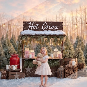 May include: A young girl stands in front of a rustic hot cocoa stand in a snowy forest setting. The stand features a sign that reads "Hot Cocoa", jars of candy, and string lights. She holds a teddy bear, wearing a white dress.