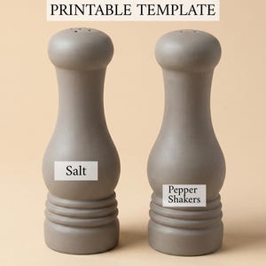 Op de afbeelding: Twee matgrijze zout- en peperstrooiers met een glad, gebogen silhouet en een geribbelde voet. De linker strooier heeft een wit label met de tekst Salt, de rechter met Pepper Shakers, en de tekst bovenaan luidt PRINTABLE TEMPLATE.