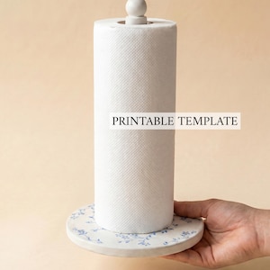 Pode incluir: Um rolo de papel toalha branco sobre uma base circular com um padrão floral azul e um pequeno puxador branco no topo. O texto PRINTABLE TEMPLATE é visível no centro do rolo de papel toalha branco.