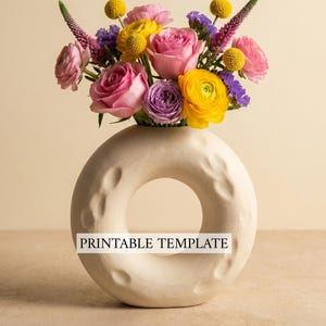 May include: A cream-colored, donut-shaped vase holds a vibrant bouquet of flowers. The arrangement includes pink roses, yellow ranunculus, purple statice, and yellow billy buttons. The vase has a textured surface and the text "PRINTABLE TEMPLATE" is displayed on the front.