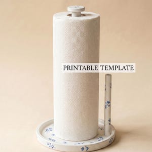Könnte beinhalten: Ein weißer Papierrollenhalter mit blauem Blumenmuster auf dem runden Sockel und dem Seitenstab, ausgestattet mit einer zentralen Spindel und einem flachen Knopf. Der Text PRINTABLE TEMPLATE steht in schwarzen Buchstaben auf der Papierrolle.