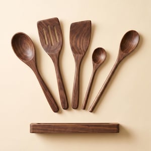 May include: A set of five wooden kitchen utensils, including spoons and spatulas, arranged on a light beige surface. The utensils are made of dark brown wood and include a slotted spatula, a flat spatula, and three different sized spoons. A wooden utensil holder is at the bottom.