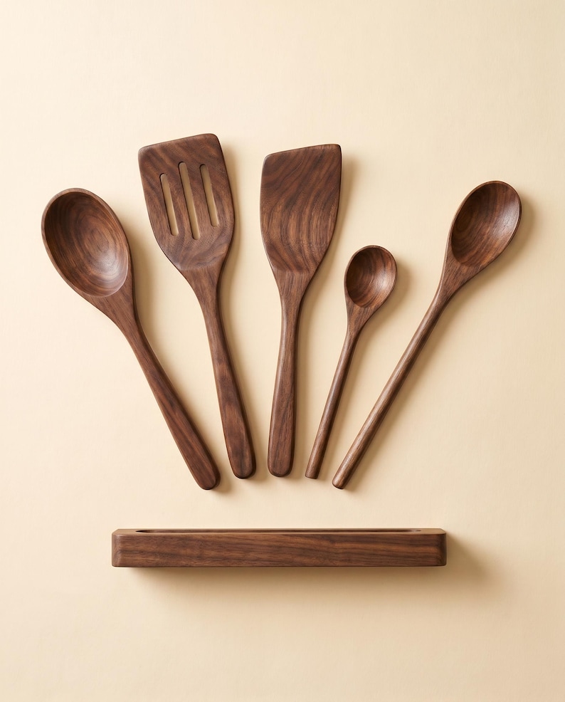 May include: A set of five wooden kitchen utensils, including spoons and spatulas, arranged on a light beige surface. The utensils are made of dark brown wood and include a slotted spatula, a flat spatula, and three different sized spoons. A wooden utensil holder is at the bottom.