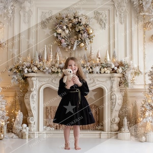 May include: A young person in a black dress holding a teddy bear stands in front of a decorated fireplace. The fireplace is adorned with a garland, ornaments, and miniature Christmas trees. Two lit Christmas trees flank the fireplace, creating a festive holiday scene.