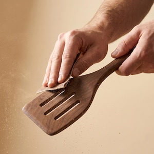 May include: A wooden spatula with a slotted design is being sanded. The spatula is a rich brown color and has a long handle. The image shows the process of woodworking, with fine particles of wood dust visible.