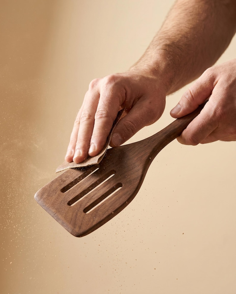 May include: A wooden spatula with a slotted design is being sanded. The spatula is a rich brown color and has a long handle. The image shows the process of woodworking, with fine particles of wood dust visible.