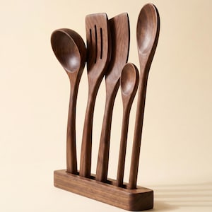 May include: A set of five wooden kitchen utensils, including a ladle, slotted spatula, solid spatula, small spoon, and large spoon, displayed in a wooden holder. The utensils are made of dark brown wood and have a smooth finish. The holder is rectangular.