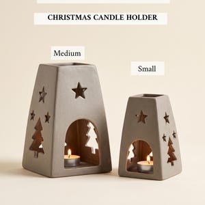 Op de afbeelding: Een printbaar sjabloon voor een kerstkaarsenhouder met twee grijze, keramiek-achtige kaarsenhouders in medium en kleine maten. Elke houder heeft een taps toelopende vorm met uitsparingen van sterren en dennenbomen, met een brandend waxinelichtje.