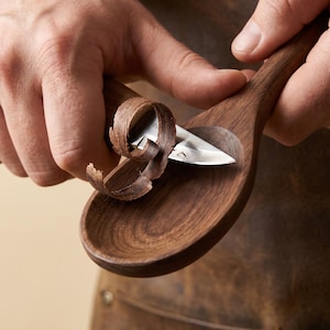 May include: A craftsman carving a wooden spoon with a small, curved knife. The spoon is made of dark brown wood, and the knife is removing thin shavings. The artisan is wearing a brown leather apron.