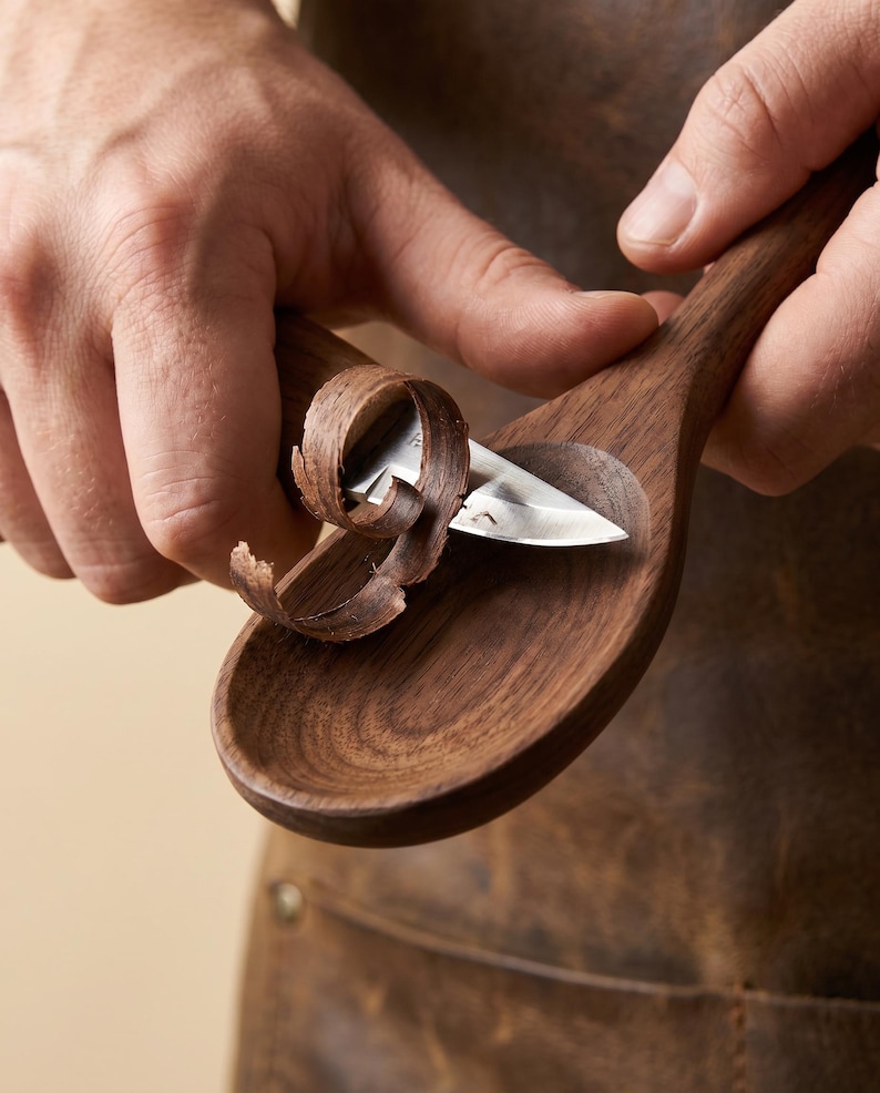 May include: A craftsman carving a wooden spoon with a small, curved knife. The spoon is made of dark brown wood, and the knife is removing thin shavings. The artisan is wearing a brown leather apron.