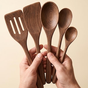 May include: A set of five wooden kitchen utensils, including a slotted spatula, solid spatula, and three spoons of varying sizes. The utensils are made of dark brown wood and held in two hands against a neutral background.