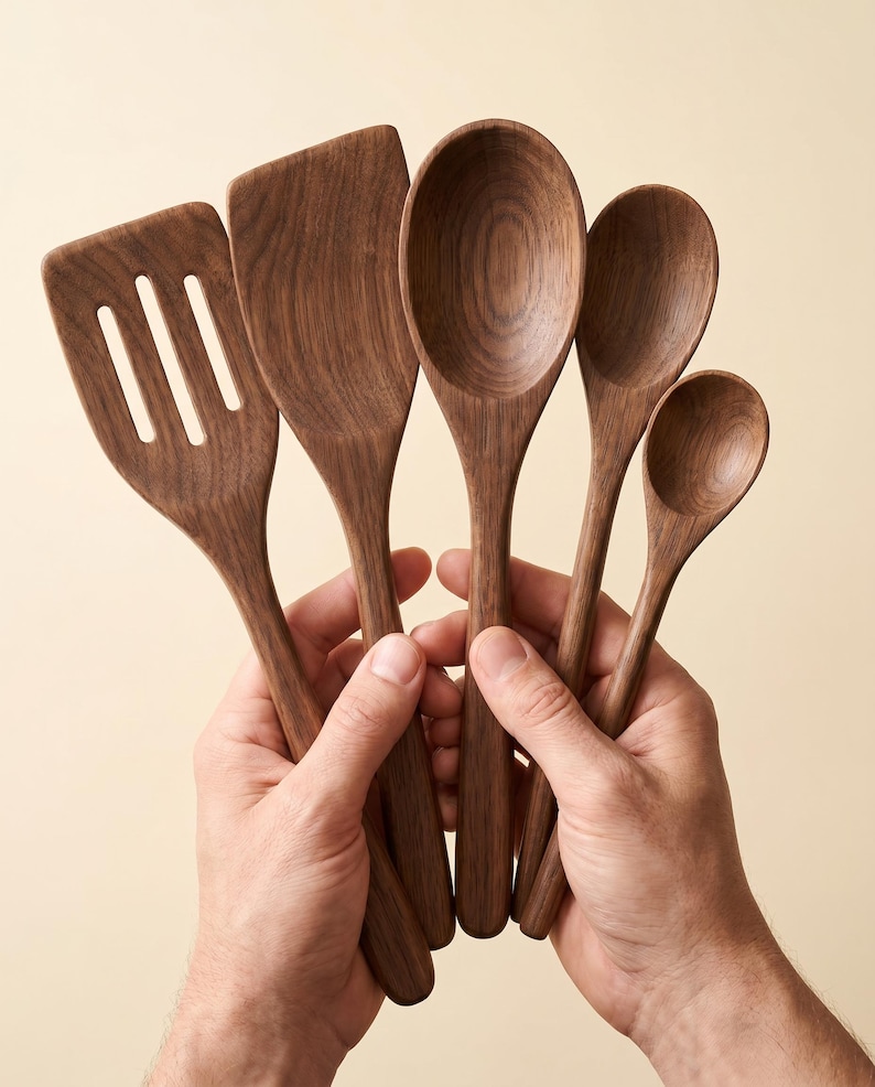 May include: A set of five wooden kitchen utensils, including a slotted spatula, solid spatula, and three spoons of varying sizes. The utensils are made of dark brown wood and held in two hands against a neutral background.