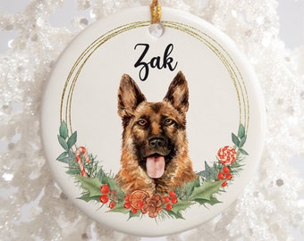 May include: A round, white ceramic ornament featuring a German Shepherd dog portrait with the name "Zak" in black script. The dog is surrounded by a festive wreath of holly, berries, and candy canes, with a gold string for hanging.