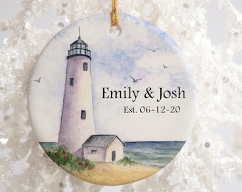 Könnte beinhalten: Ein weißer Keramik-Anhänger mit einem Aquarellbild eines Leuchtturms, eines kleinen Hauses und des Meeres. Der Anhänger trägt den Text "Emily & Josh Est. 06-12-20".