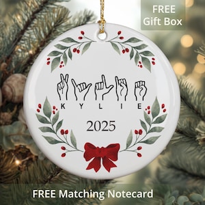 Puede incluir: Adorno navideño redondo de cerámica blanca con una cuerda dorada para colgar. El adorno presenta el nombre "KYLIE" en lenguaje de señas americano, el año "2025", un lazo rojo y un diseño de corona floral verde y roja.