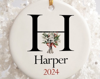 Puede incluir: Adorno navideño de cerámica blanca con una letra H negra y una cinta roja. El adorno está decorado con un ramo de bayas blancas y hojas verdes. El nombre "Harper" y el año "2024" están escritos en rojo debajo de la letra.