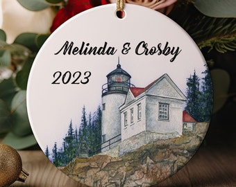 Könnte beinhalten: Ein weißer Keramik-Anhänger mit einem Aquarellbild eines Leuchtturms auf einer felsigen Klippe. Der Anhänger ist personalisiert mit den Namen "Melinda & Crosby" und dem Jahr "2023".