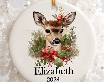 May include: A white ceramic Christmas ornament with a watercolor illustration of a fawn with a red and green holly wreath. The ornament is personalized with the name "Elizabeth" and the year "2024".