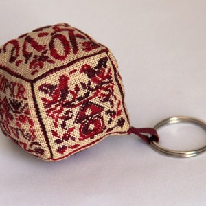 Può includere: Un portachiavi a forma di cubo ricamato beige e rosso con un anello portachiavi argentato. Il cubo presenta una varietà di disegni su ogni lato, tra cui una casa, un uccello e un fiore.