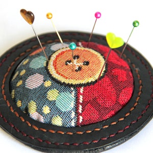 May include: A round pincushion with a colorful fabric cover and a black leather trim. The pincushion has a button design in the center and is filled with pins with colorful heads.
