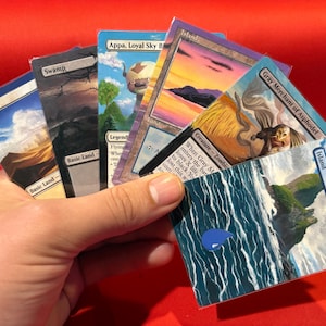 May include: A hand holding a fan of Magic: The Gathering trading cards. The cards feature various landscapes and creatures, including "Plains," "Swamp," and "Island." The cards have text such as "Gray Merchant of Asphodel" and "Appa, Loyal Sky B."