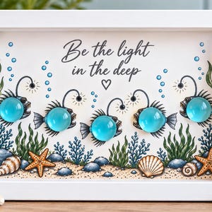May include: A white framed wall art piece featuring an underwater scene with five illustrated anglerfish, each with a bright blue glass cabochon body. The text Be the light in the deep is printed above the fish, surrounded by seaweed and seashells.