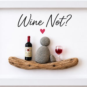 May include: A framed wall art piece featuring the text Wine Not? in black script above a red heart. Below, a miniature wine bottle, a figure made of two grey stones, and a small glass of red wine are arranged on a piece of textured driftwood.