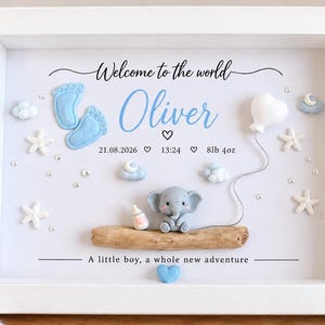 May include: A white framed shadow box featuring the text Welcome to the world Oliver, 21.08.2026, 13:24, and 8lb 4oz. The display includes blue baby footprints, a small elephant figurine, a white heart balloon, stars, clouds, and a piece of driftwood.