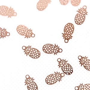 Könnte beinhalten: Roségoldfarbene Ananas-Anhänger auf weißem Untergrund. Die Anhänger sind als Ausschnitte gestaltet, die die Textur und Form der Ananas zeigen. Jeder Anhänger hat eine kleine Schlaufe zum Befestigen an Schmuck oder anderen Bastelarbeiten.